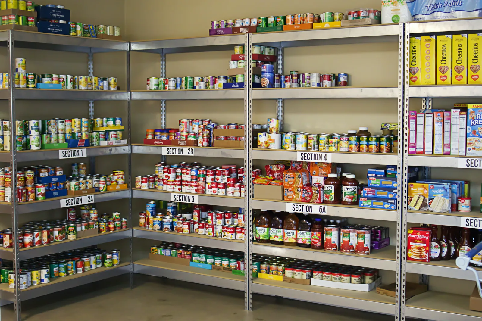Metal shelving units in a pantry stocked with canned vegetables, soups, pasta sauce, juice and boxes of cereal, each shelf marked with a numbered section label