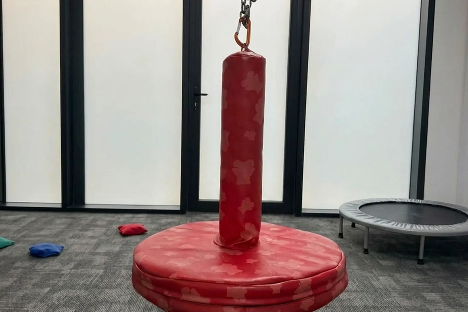 A red padded platform swing hangs from a chain in an empty therapy room with frosted windows, a small trampoline and bean bags on a gray carpet