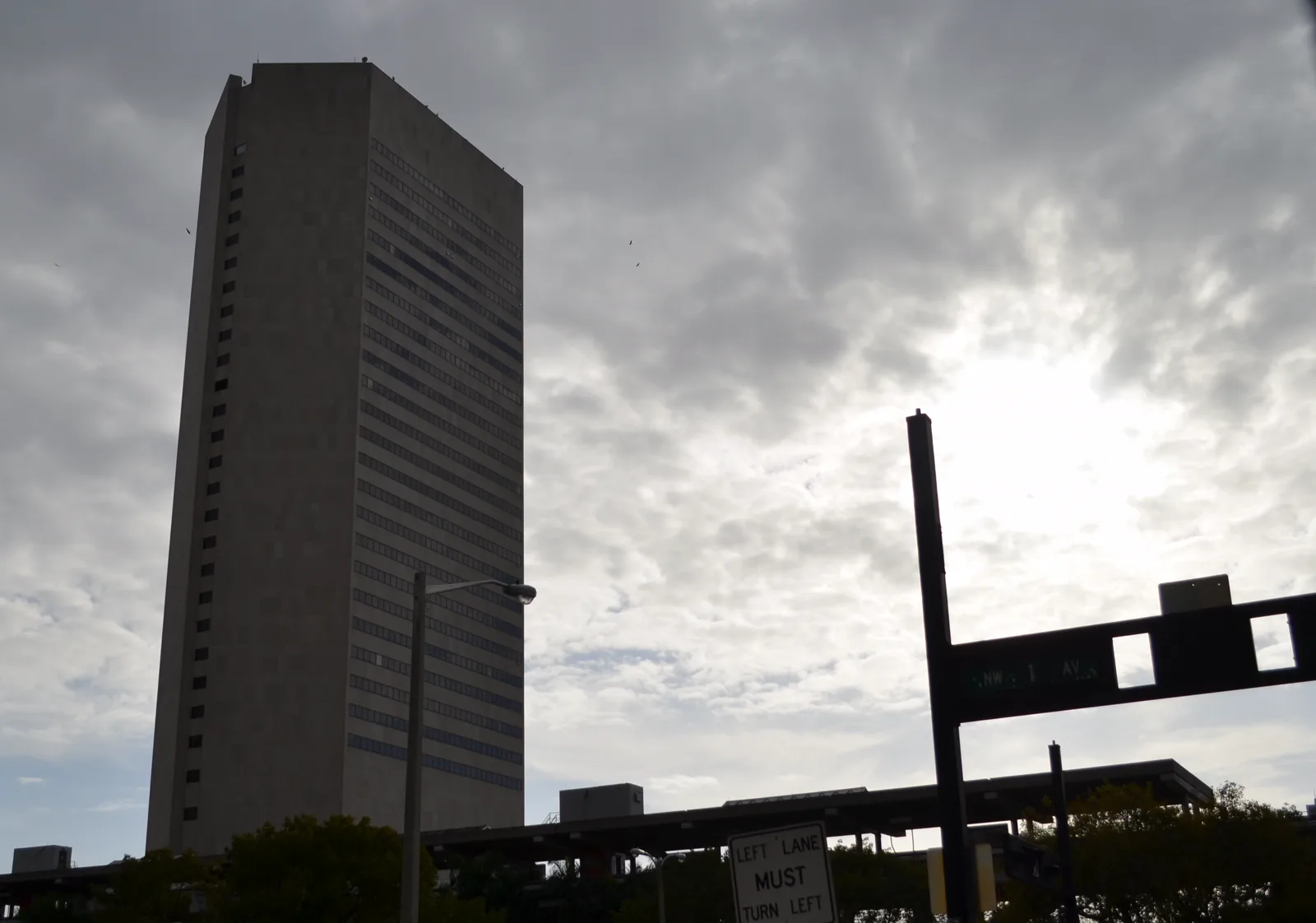 A tall rectangular office tower with rows of narrow windows under a cloudy gray sky, with trees, a light pole, a traffic sign and an elevated rail line in the foreground