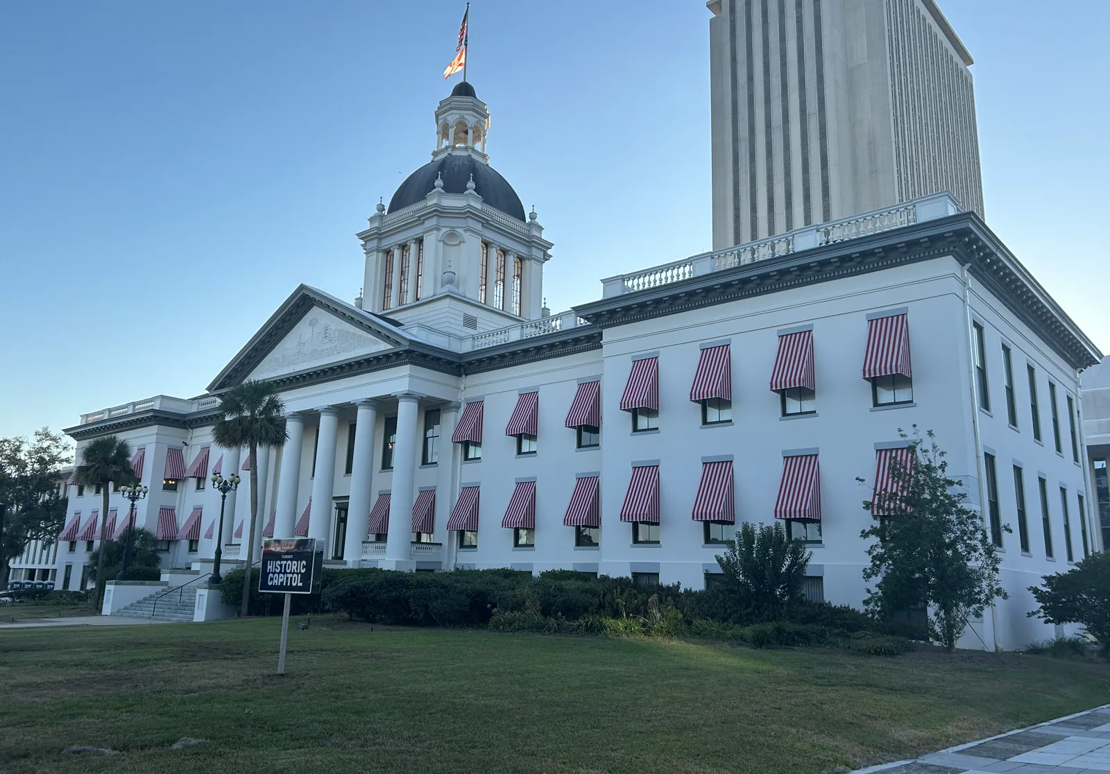 A white domed building with red and white striped awnings and a columned portico, with a tall modern office tower rising behind it under a clear sky