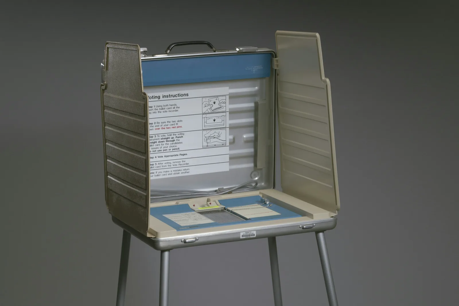 A portable aluminum voting booth on thin metal legs with beige side panels, a printed sheet of voting instructions and a blue ballot card holder, against a plain gray background