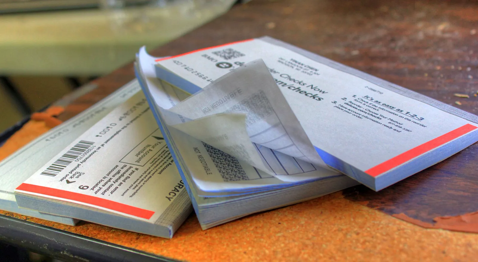 Three paper checkbooks lie fanned out on a worn wooden table, one opened with its pages curling back, a barcode printed on the cover of another