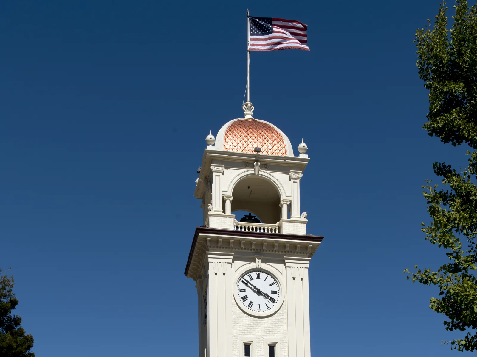The top of a white clock tower with a salmon-colored tiled dome and an open belfry, an American flag flying above it, against a deep blue sky with a tree at the right edge