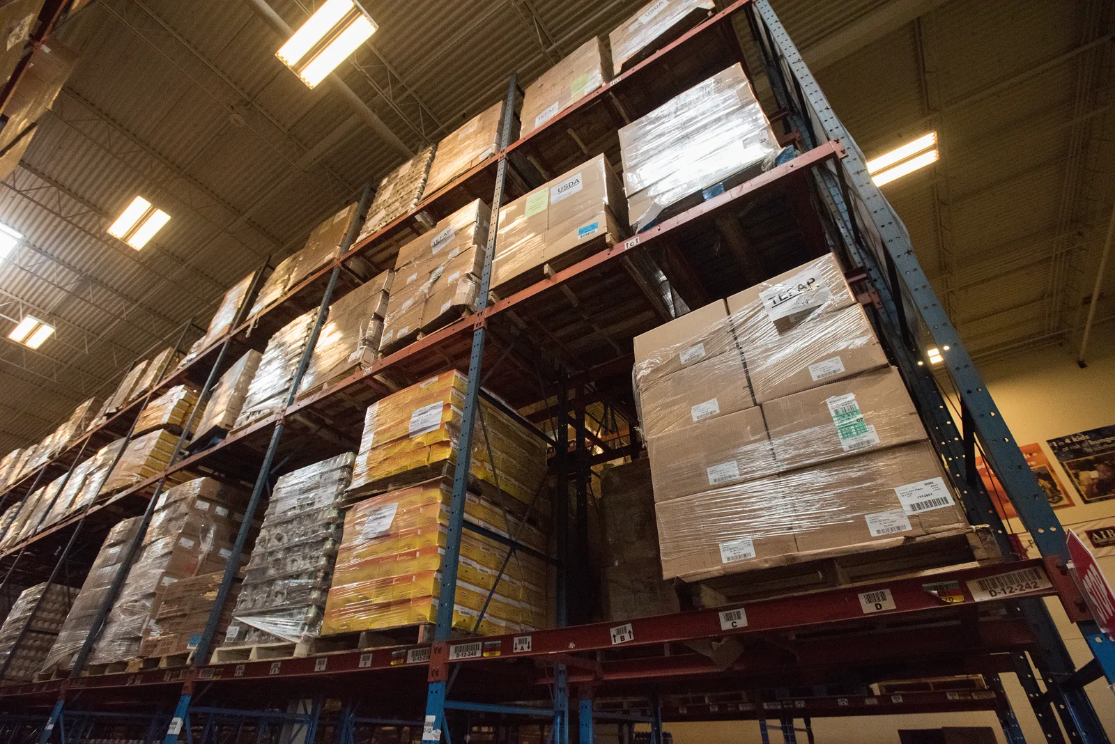 Tall red and blue steel racks in a warehouse stacked to the ceiling with shrink-wrapped pallets of boxed food, seen from below under rows of fluorescent lights