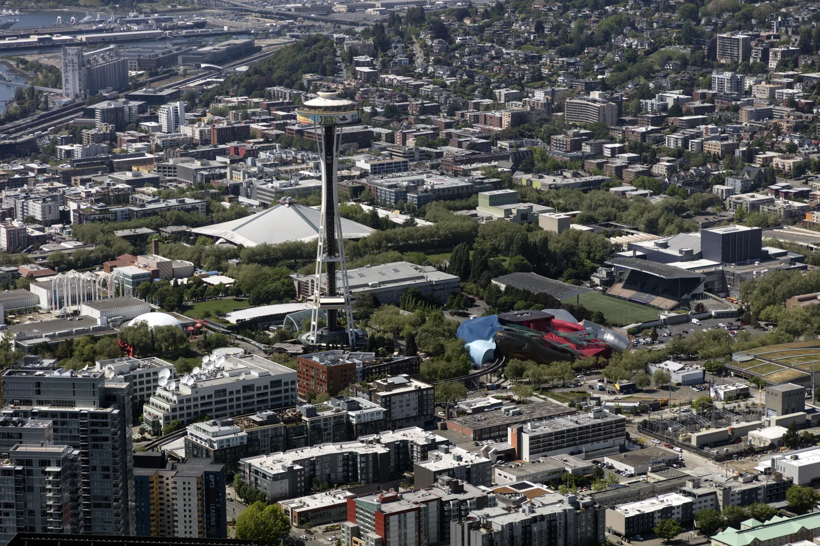 An aerial view of a city neighborhood with a tall, slender observation tower at the center, a white peaked arena roof behind it, a curving red and blue museum building and a sports field to the right, and apartment blocks in the foreground