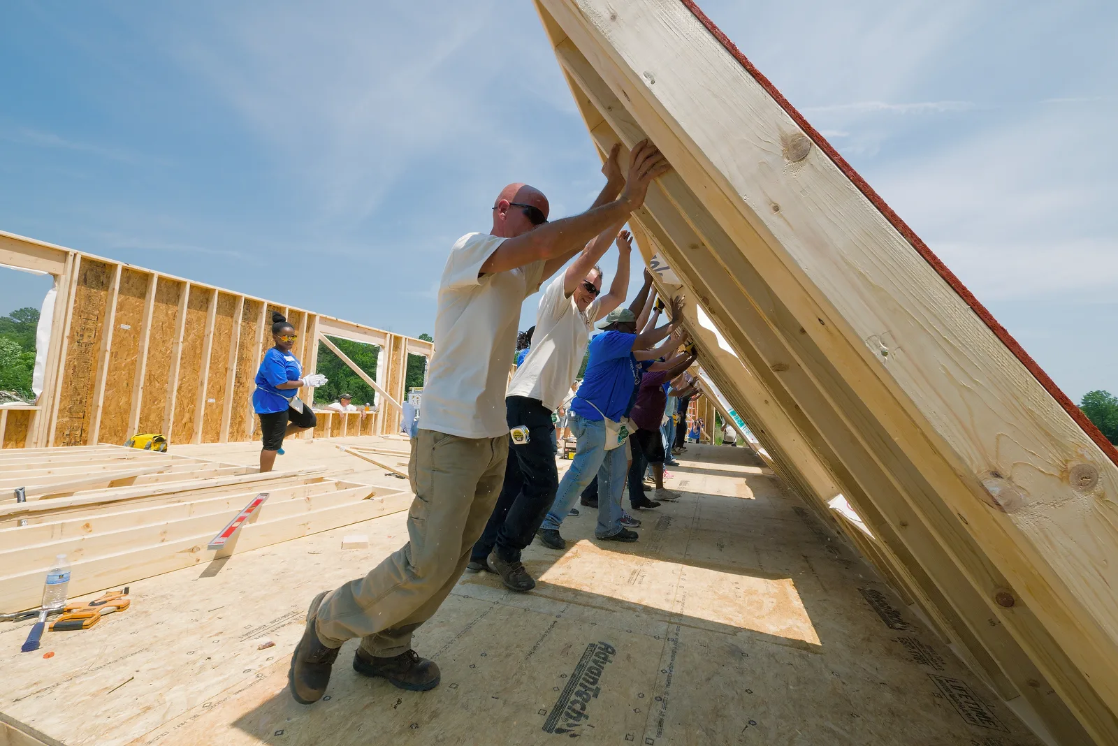 A row of volunteers in work clothes and boots push a tall wooden wall frame upright on the plywood floor of a new house, with other framed walls behind them under a blue sky