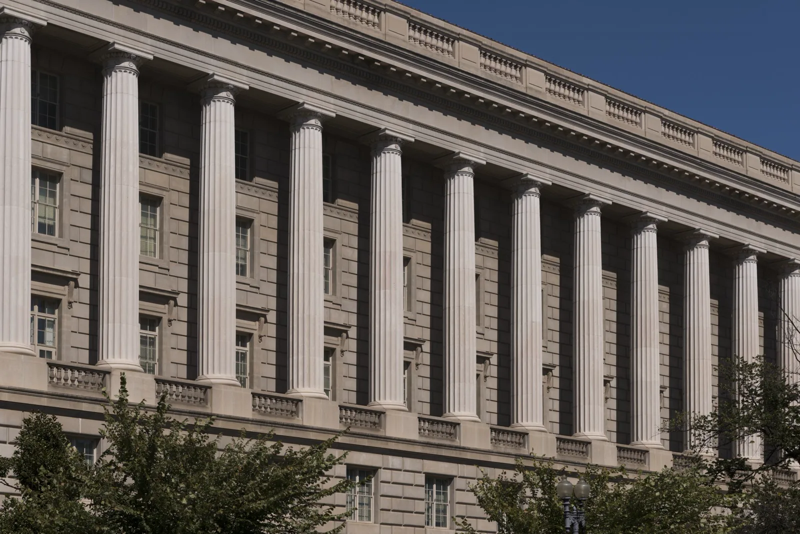 A row of tall fluted stone columns along the upper floors of a long gray government building, seen at an angle against a deep blue sky, with treetops along the bottom