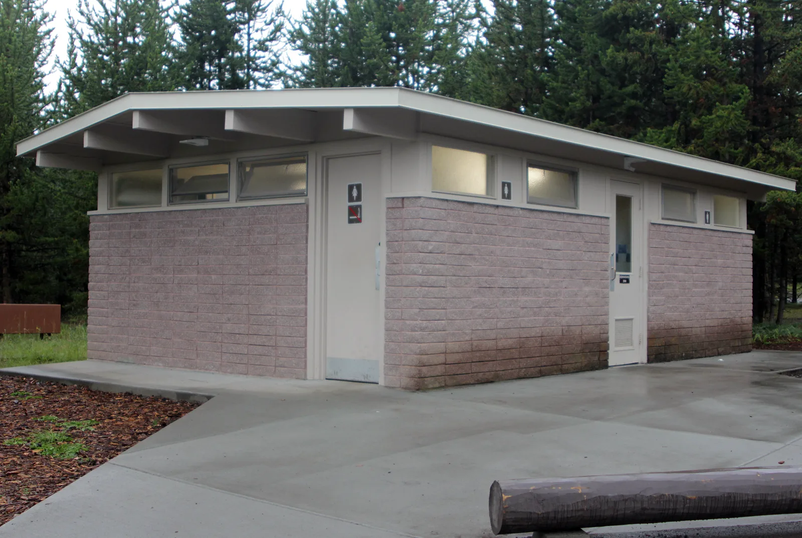 A small single-story restroom building with pinkish block walls, a flat overhanging roof, high windows and two doors marked with restroom signs, on a concrete apron among tall evergreen trees