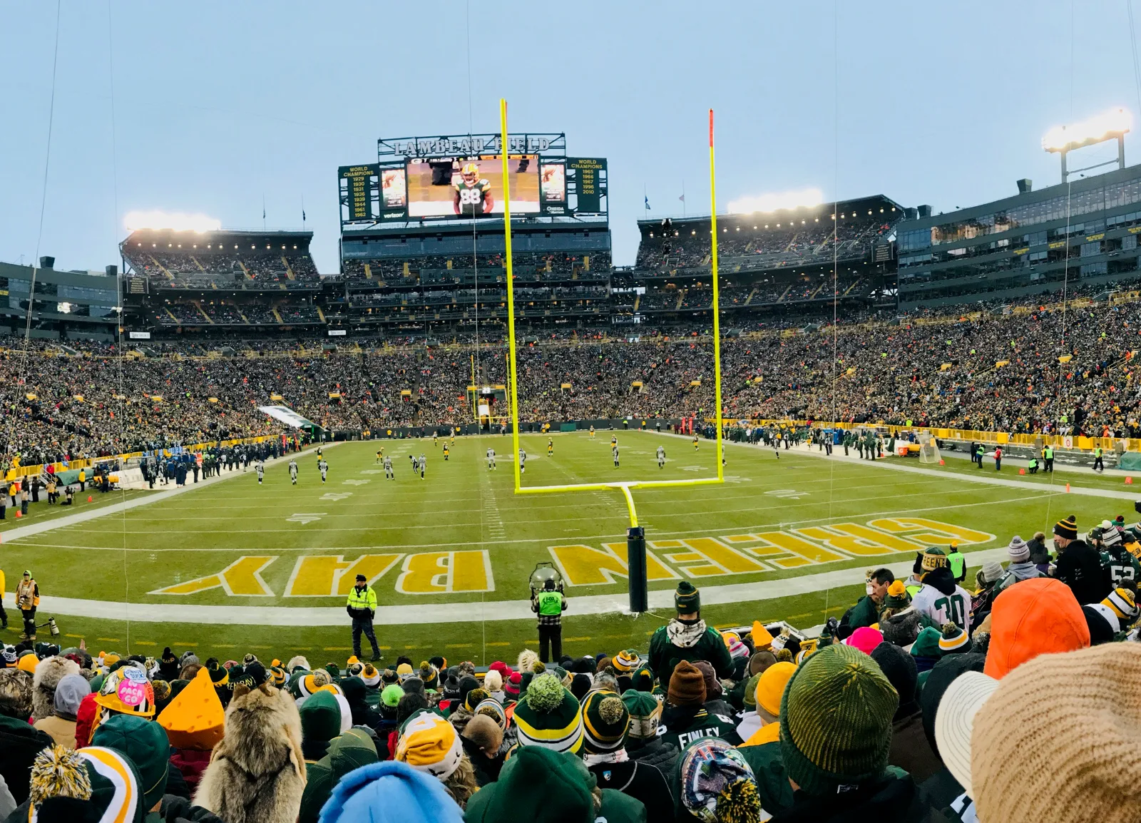 Lambeau Field seen from the lower stands behind an end zone, with GREEN BAY painted on the grass, a yellow goalpost, players on the field and a full crowd in green and gold hats in the foreground