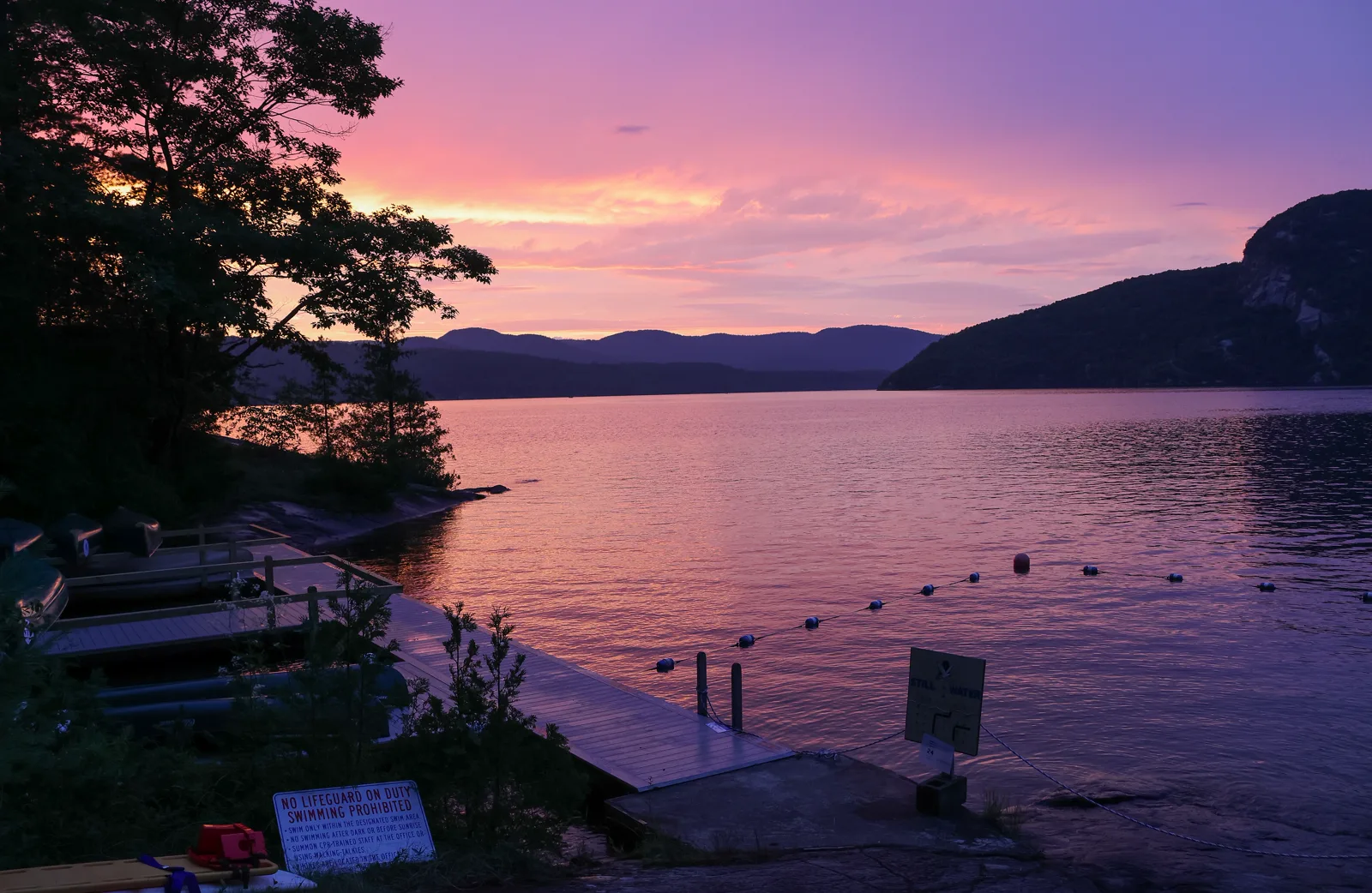 Canoes on racks beside a wooden dock and a roped swimming area on a lake at sunset, with wooded mountains behind