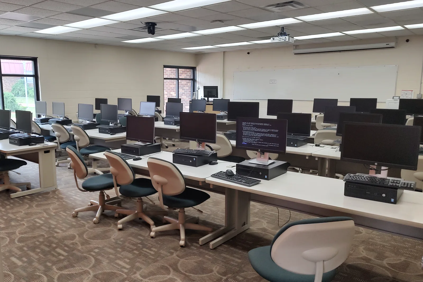 An empty classroom with rows of white desks holding desktop computers, monitors and keyboards, green and beige office chairs, a whiteboard on the far wall and windows on the left