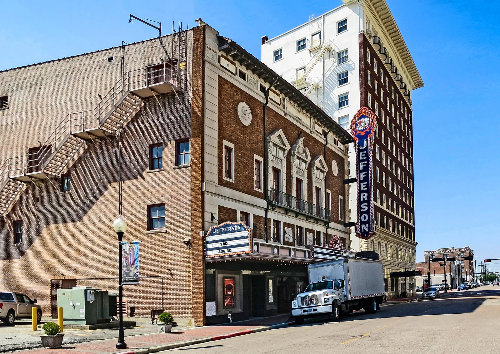 A brick theater with a lit marquee and a tall vertical Jefferson sign on a downtown street, a box truck parked in front and a white high-rise behind it under a clear blue sky