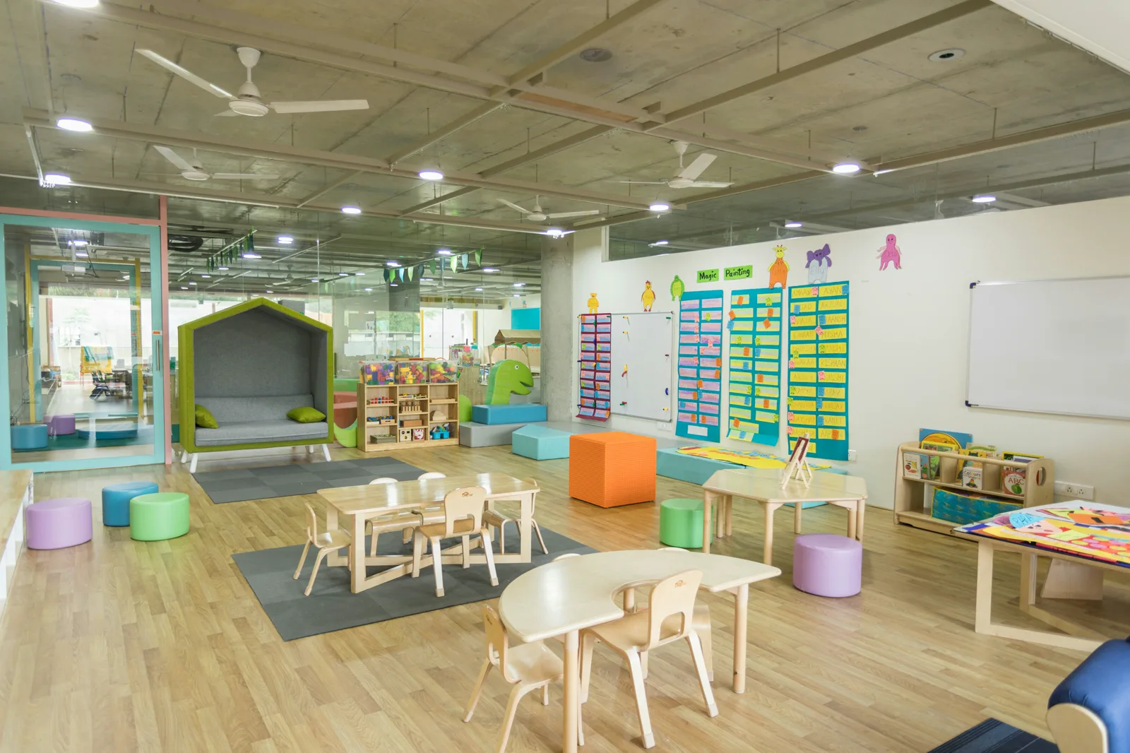 An empty early childhood classroom with low wooden tables and child-size chairs, round cushioned stools, a reading nook shaped like a small house and charts on the wall