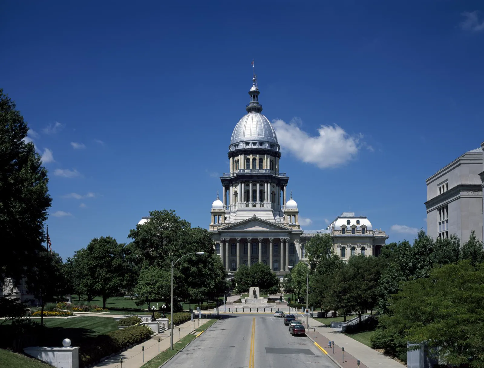 A domed stone capitol building with a columned portico and a flag on top, seen down a street lined with trees, lawns and parked cars under a deep blue sky