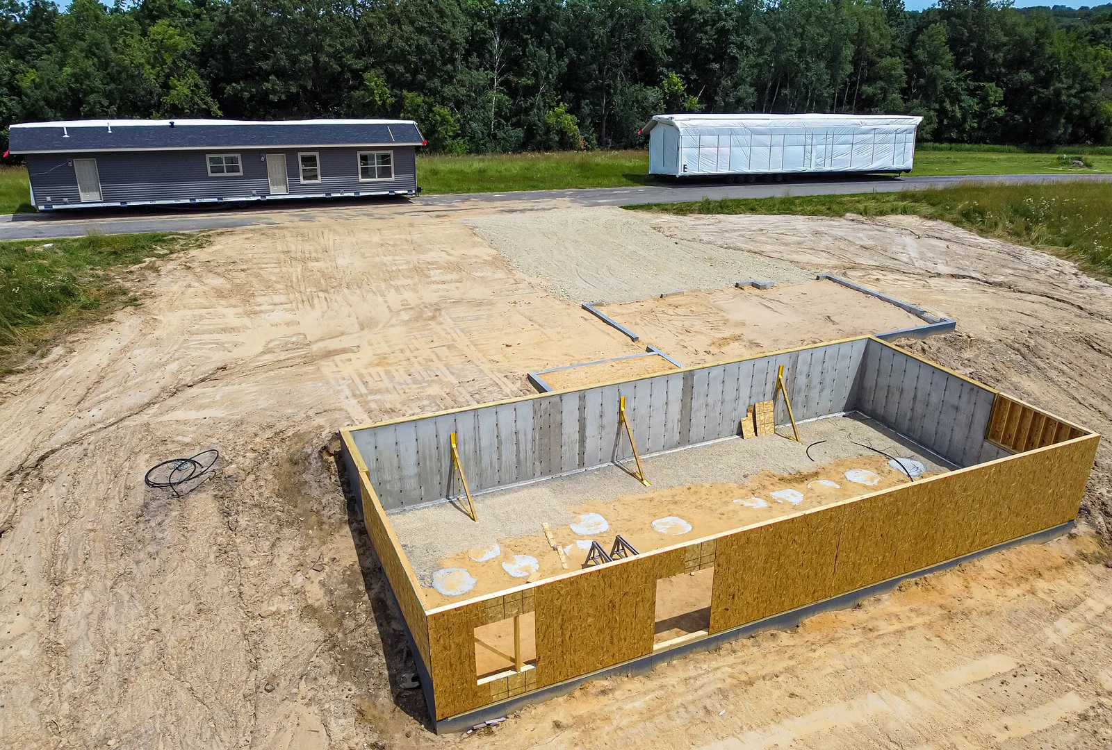 Aerial view of a new foundation of concrete block and wooden walls on a cleared lot, with two sections of a prefabricated house waiting on a road behind it, one wrapped in white plastic