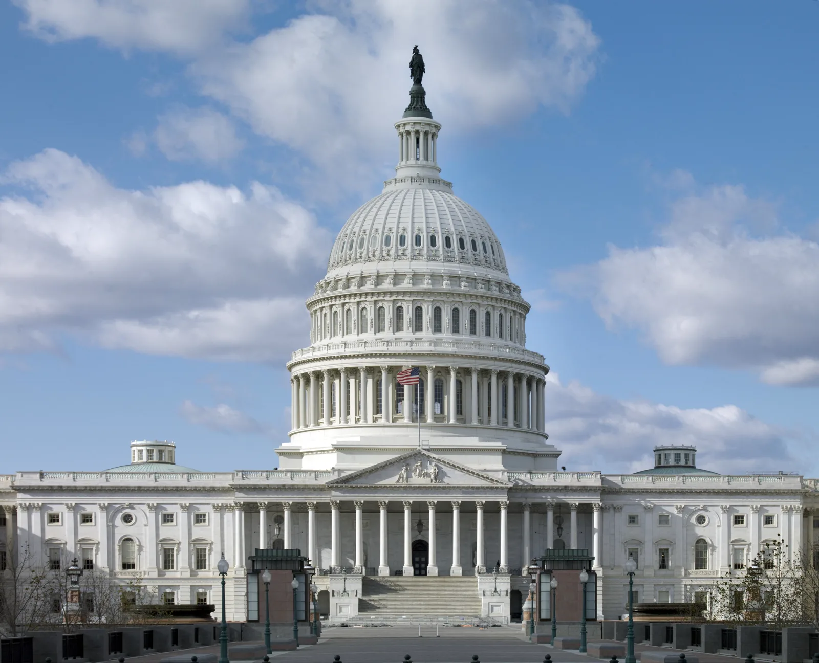 The white domed United States Capitol seen head on from the east across an empty plaza, with a flag flying below the dome and white clouds in a blue sky