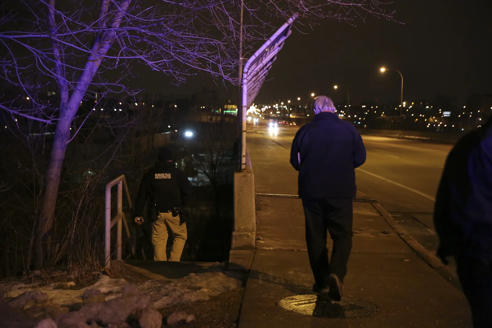 At night, a man in a dark jacket walks away along a bridge sidewalk while a police outreach officer, seen from behind, steps down a path beside it toward the brush below