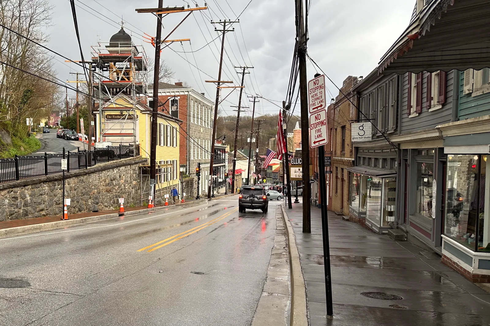 A wet two-lane street climbing between old stone, brick and clapboard buildings, with a yellow building under scaffolding on the left, shop windows on the right, utility poles and American flags, under a gray sky