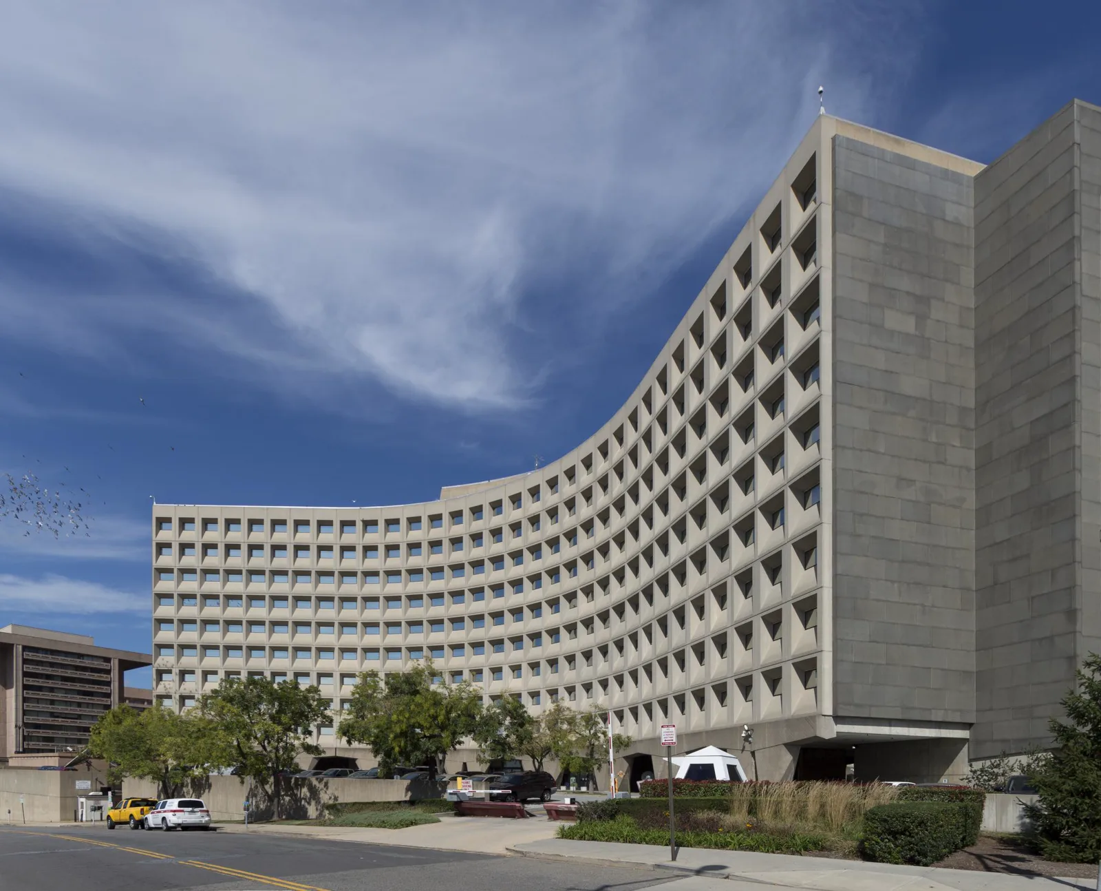 A curved ten-story concrete office building with rows of deep square windows under a blue sky with thin clouds, trees and parked cars along the street in front