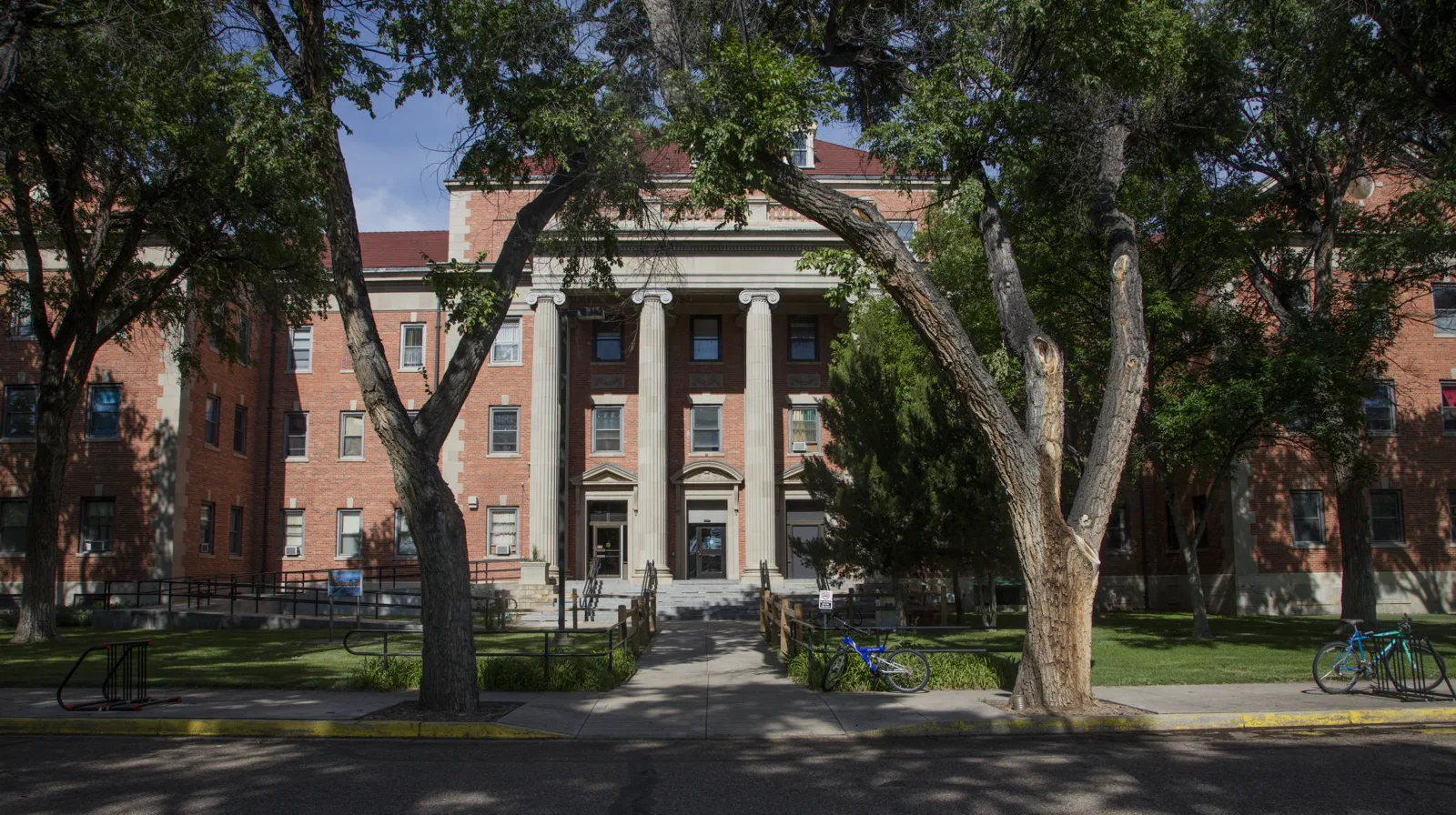 A four-story red brick building with a portico of tall white columns, seen through the trunks of large shade trees, with two bicycles parked along the walk in front