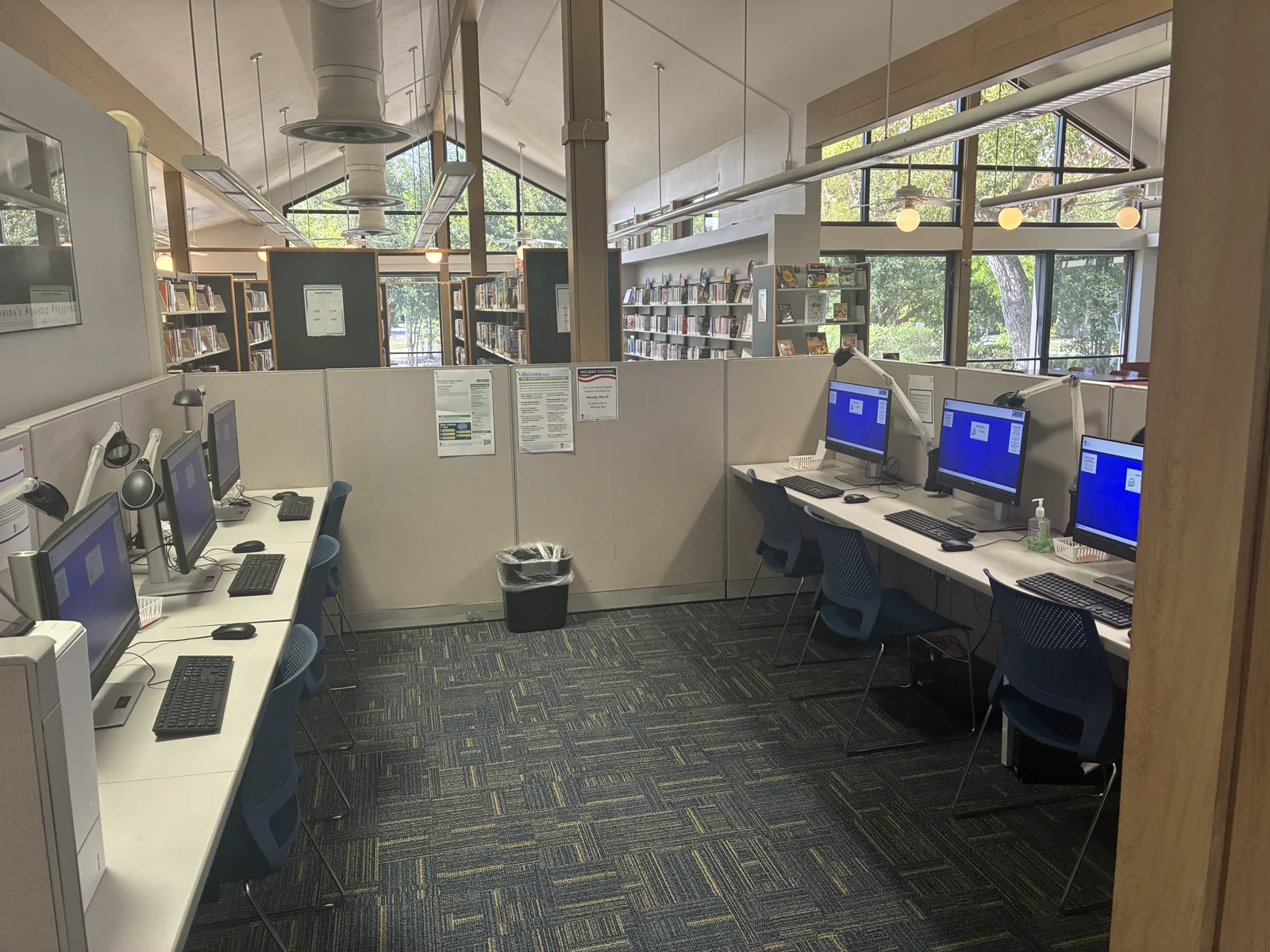 Two rows of empty computer stations with blue chairs between beige cubicle panels in a public library, with bookshelves and tall windows behind them