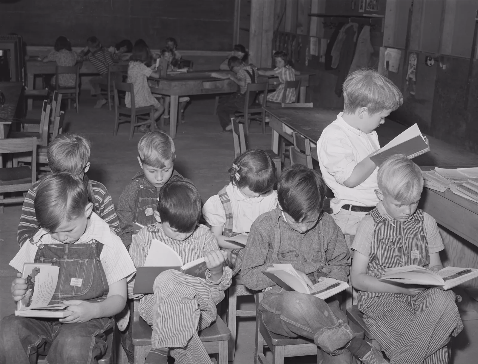 Black and white photo of a schoolroom where children in overalls sit on small wooden chairs with their heads bent over open books, while other children work at tables behind them