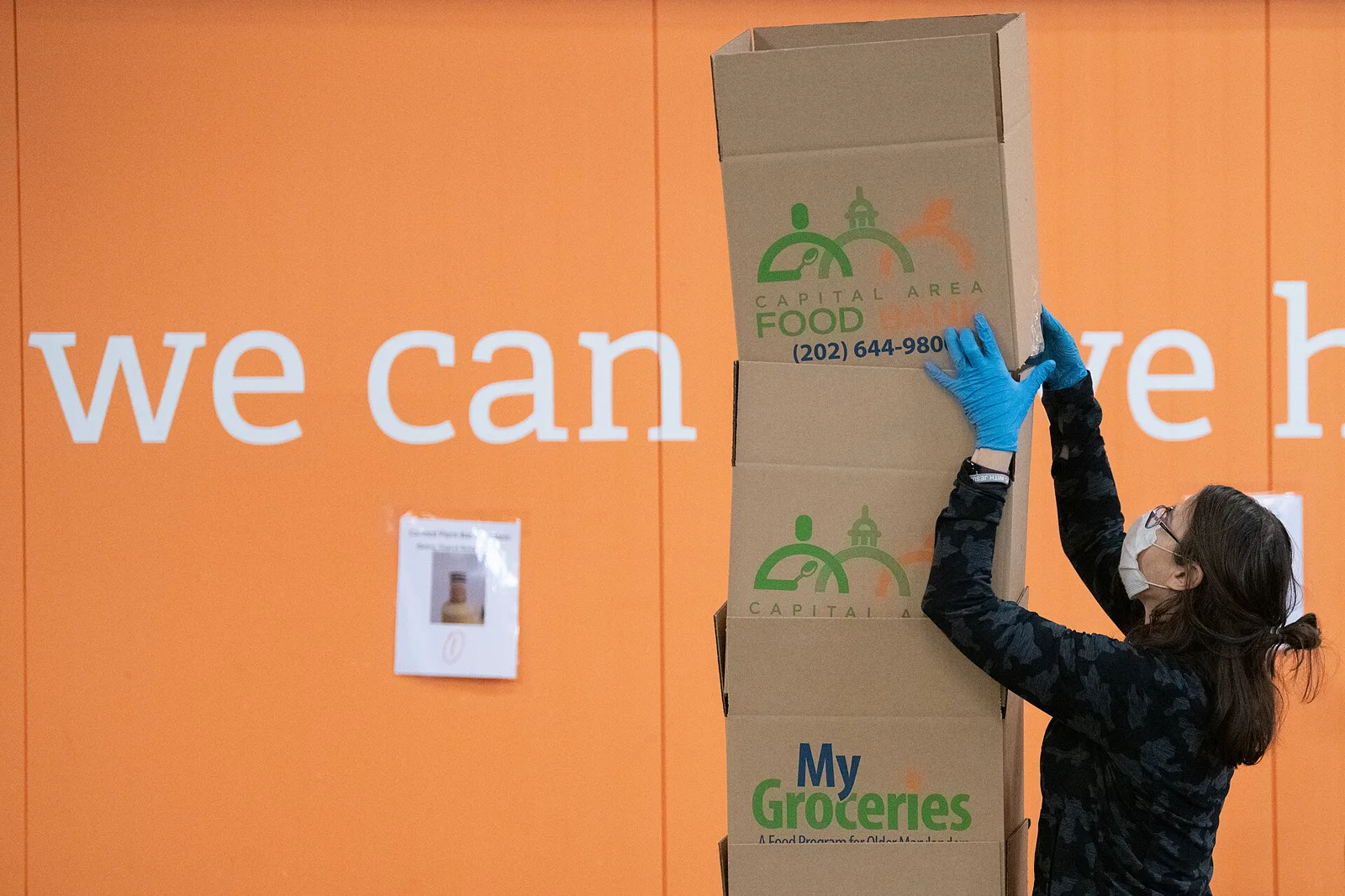 A volunteer in a mask and blue gloves reaches up to steady a tall stack of cardboard grocery boxes in front of an orange warehouse wall