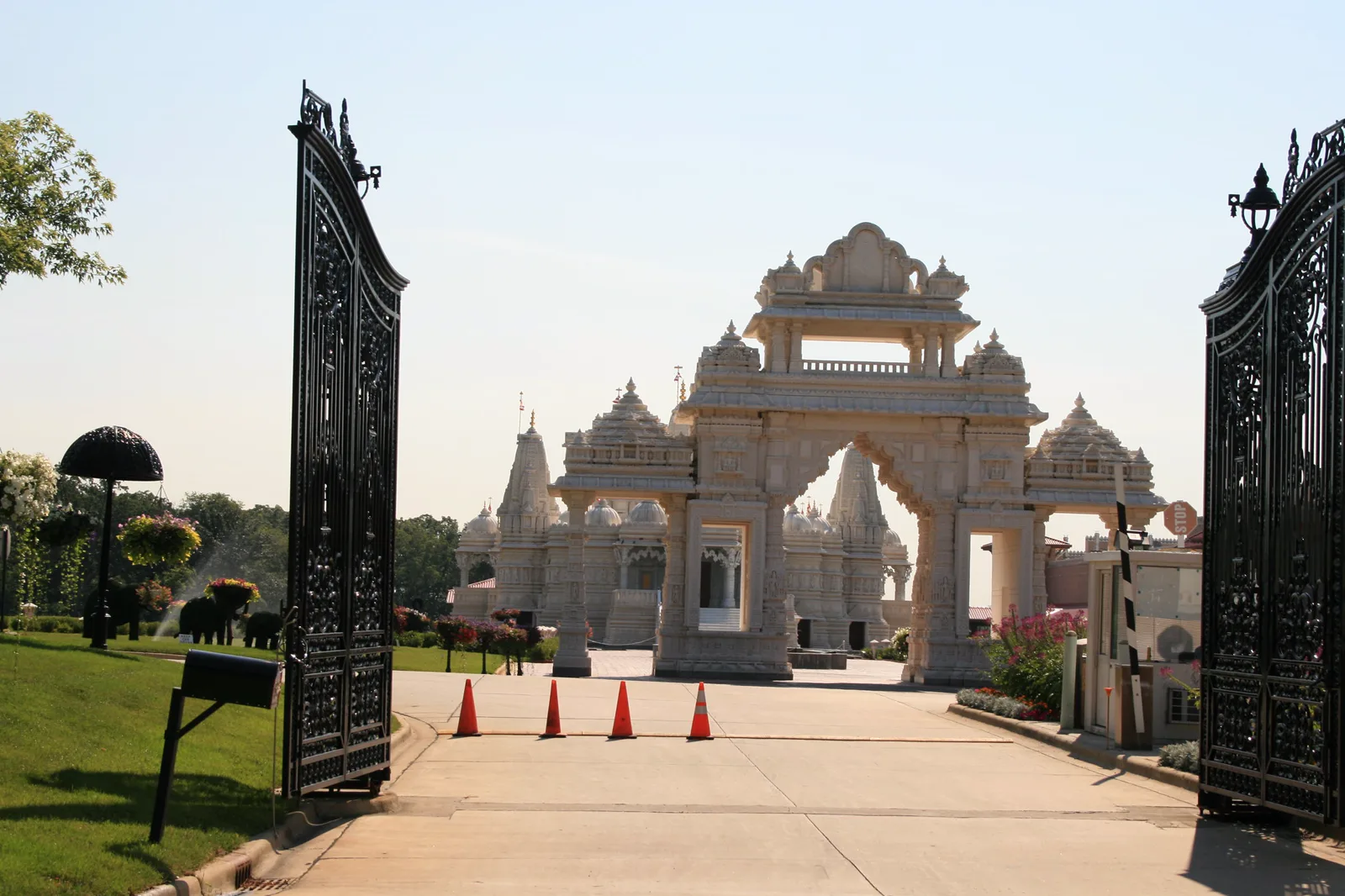 Tall black iron gates stand open on a driveway lined with orange traffic cones, leading to a white carved stone gateway and temple domes under a clear sky