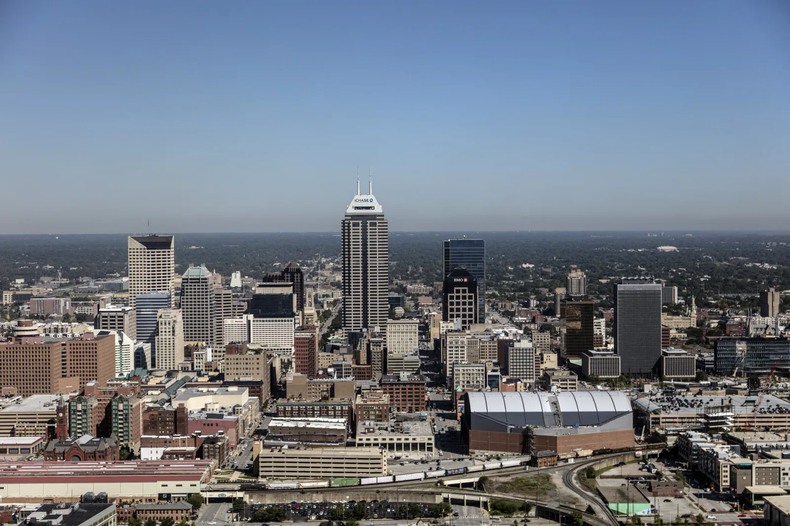 An aerial view of downtown Indianapolis on a clear day, with a tall office tower topped by two spires at the center, other high-rise buildings around it, a domed convention hall and railroad tracks in the foreground and tree-covered neighborhoods stretching to the horizon