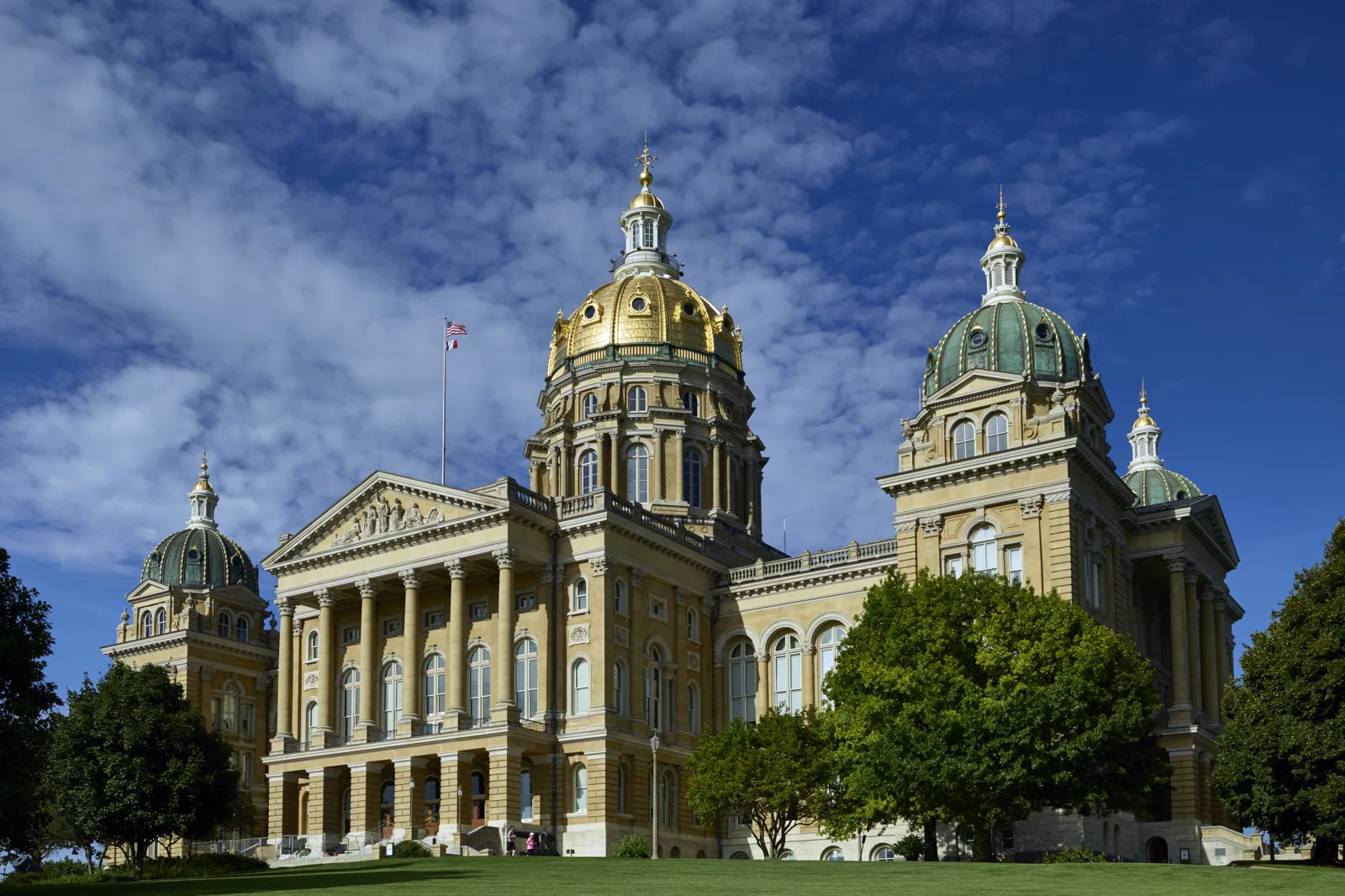 The Iowa State Capitol, a pale stone building with a columned portico, a large gilded central dome and smaller green domes at its corners, behind a lawn and trees under a blue sky with white clouds
