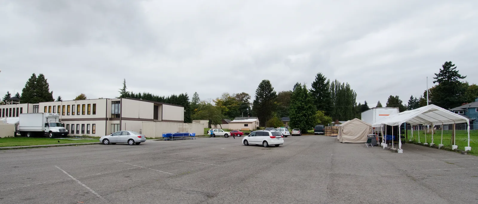 A wide parking lot under an overcast sky, with white tents, shopping carts and a box truck beside a long two-story building and tall evergreen trees; one person walks in the distance