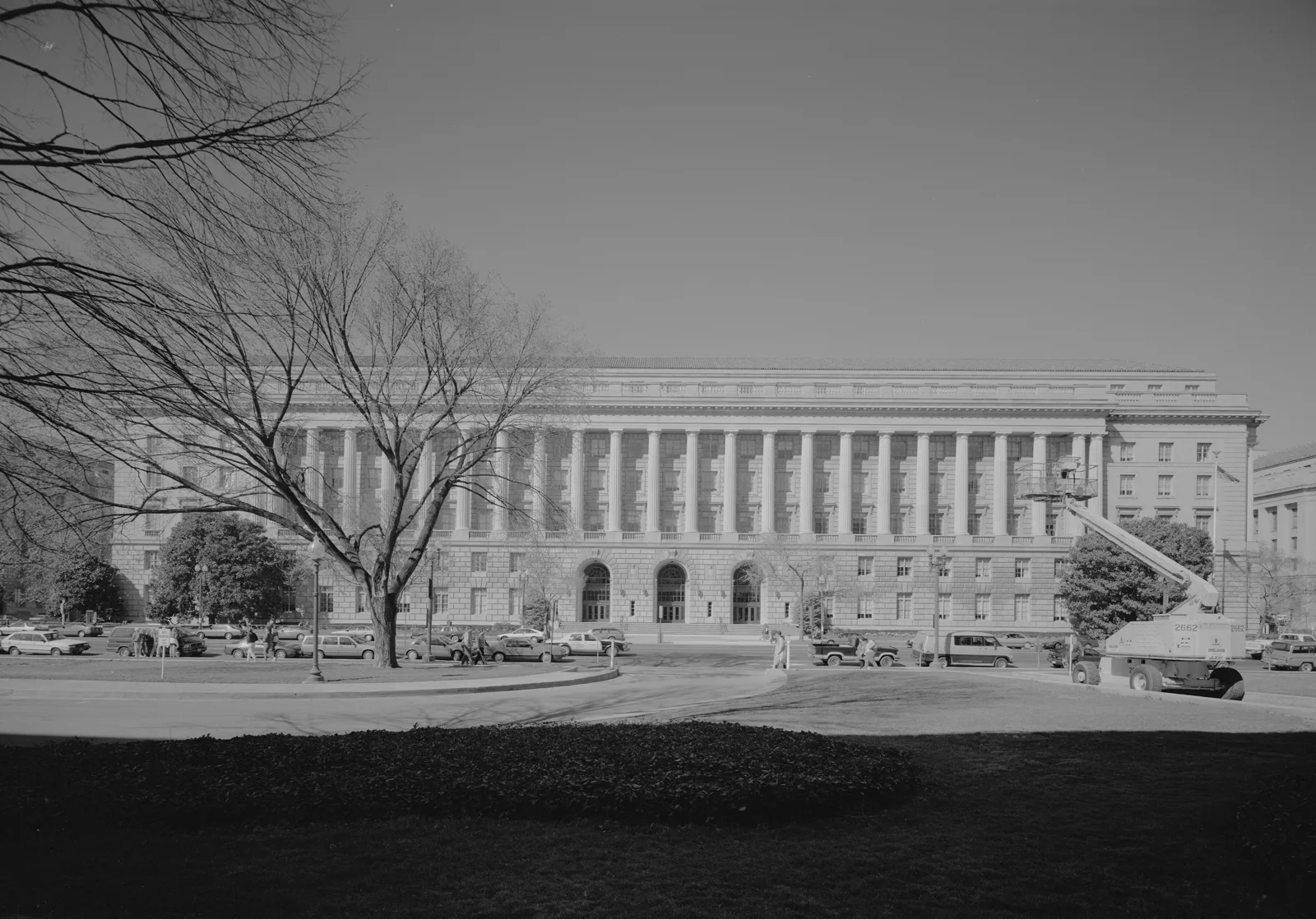 Black and white photograph of a long stone building with a row of tall columns above three arched entrances, seen across a lawn and a street lined with parked cars, with bare trees on the left and a lift truck on the right