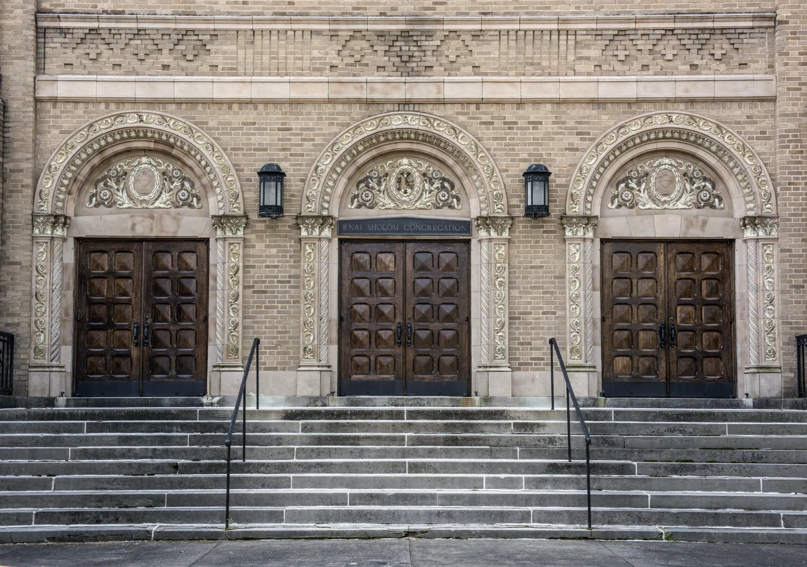 Three tall closed wooden double doors under carved stone arches in a tan brick synagogue front, with black lanterns between them and a wide flight of gray steps with two iron handrails