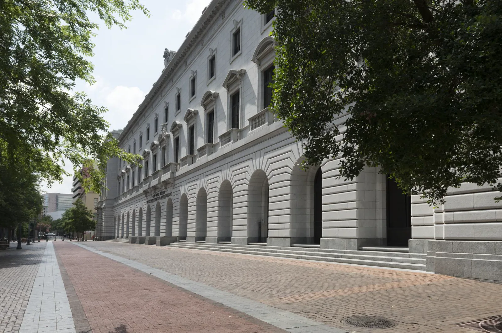 A long white stone courthouse with a row of arched openings on the ground floor and tall windows above, seen down a brick-paved sidewalk under trees