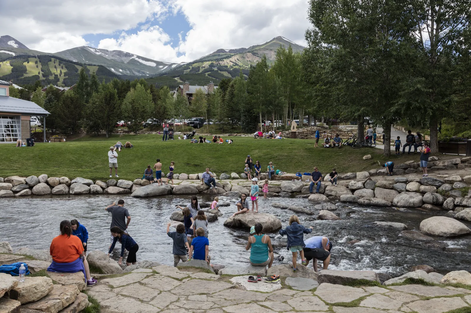 Families and children seen from behind and at a distance sit on boulders and wade in a shallow mountain river beside a stone path and a lawn, with pine trees, picnickers and forested peaks under a cloudy sky beyond