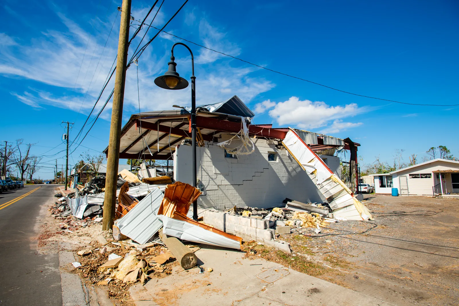 A small cinder block building with its metal roof torn open and twisted roofing and debris piled along the sidewalk of a street under a blue sky