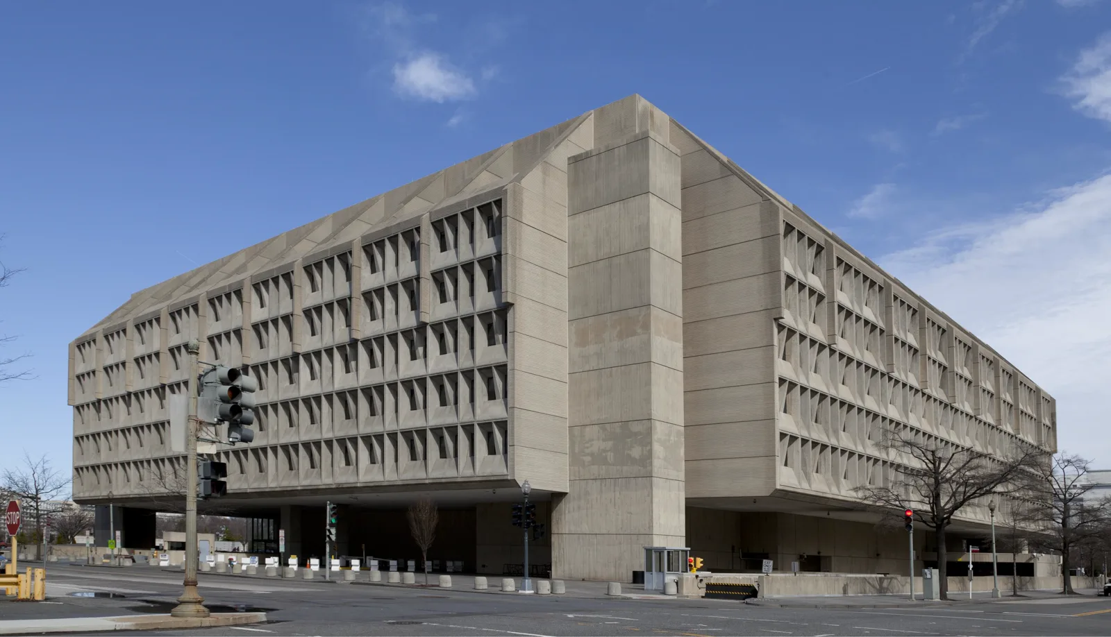 A large concrete office building with deep-set rows of windows, raised on thick columns above a street corner with traffic lights, under a blue sky