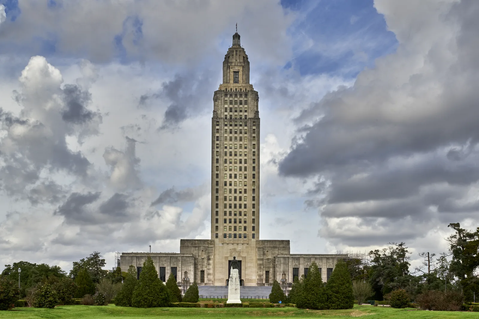 A tall art deco stone tower rising from a wide, low base under a cloudy blue sky, with a statue on a white pedestal, cone-shaped evergreens and a green lawn in front