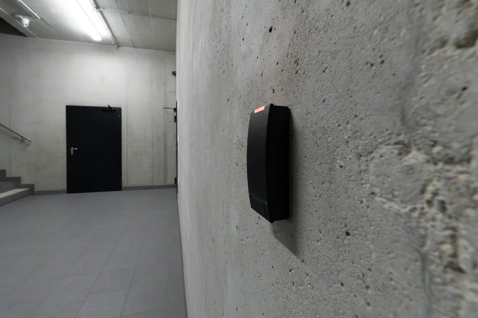 A black access card reader with a red light mounted on a rough concrete wall, beside an empty stairwell hall with a closed black door and a small dome camera on the ceiling