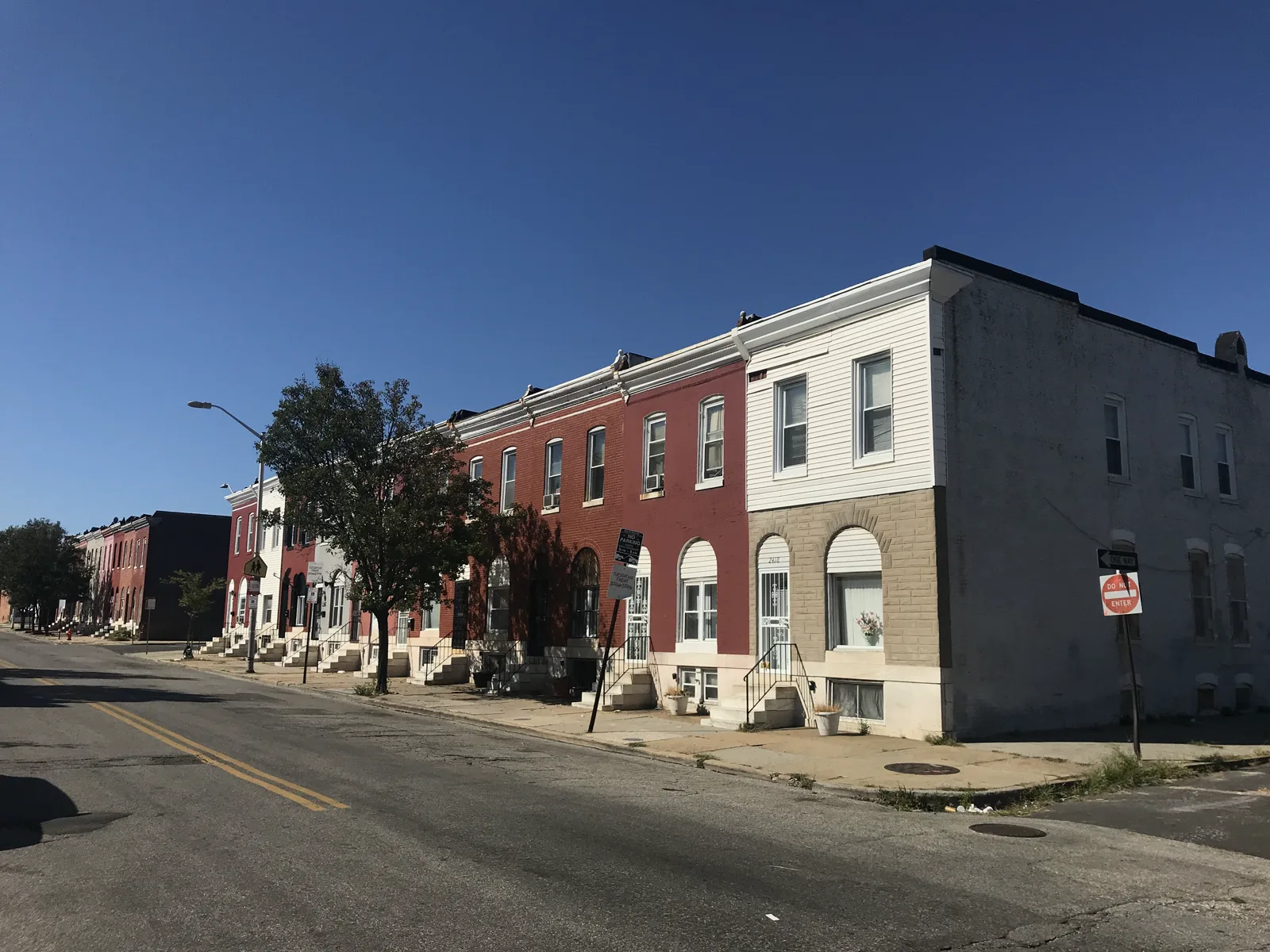 A row of two-story brick and painted rowhouses with white marble steps along an empty street under a clear blue sky, with a tree, a street lamp and a do not enter sign