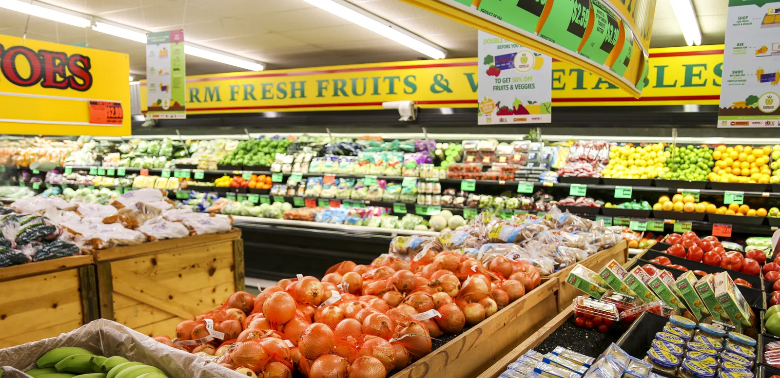 The produce section of a grocery store under a yellow sign reading Farm Fresh Fruits and Vegetables, with refrigerated shelves of greens and citrus behind wooden bins of bagged onions and potatoes and trays of tomatoes