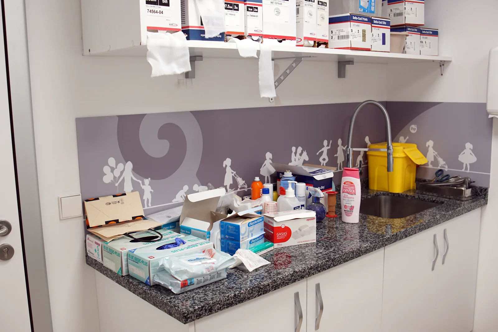 A clinic counter crowded with boxes of gloves, gauze and bottles beside a sink, with more boxes of medical supplies on a shelf above