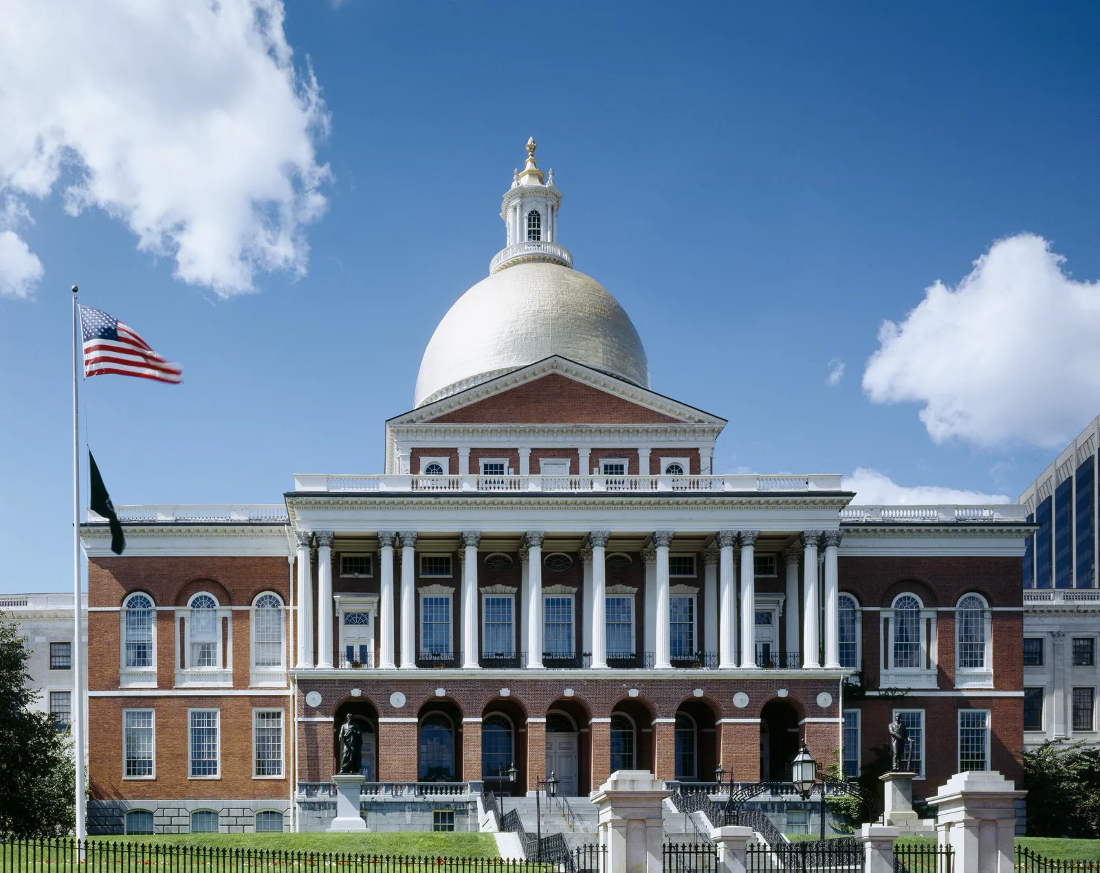 A red brick statehouse with a white columned portico and a gold dome under a blue sky with white clouds, an American flag flying on a pole at left and an iron fence along the lawn in front