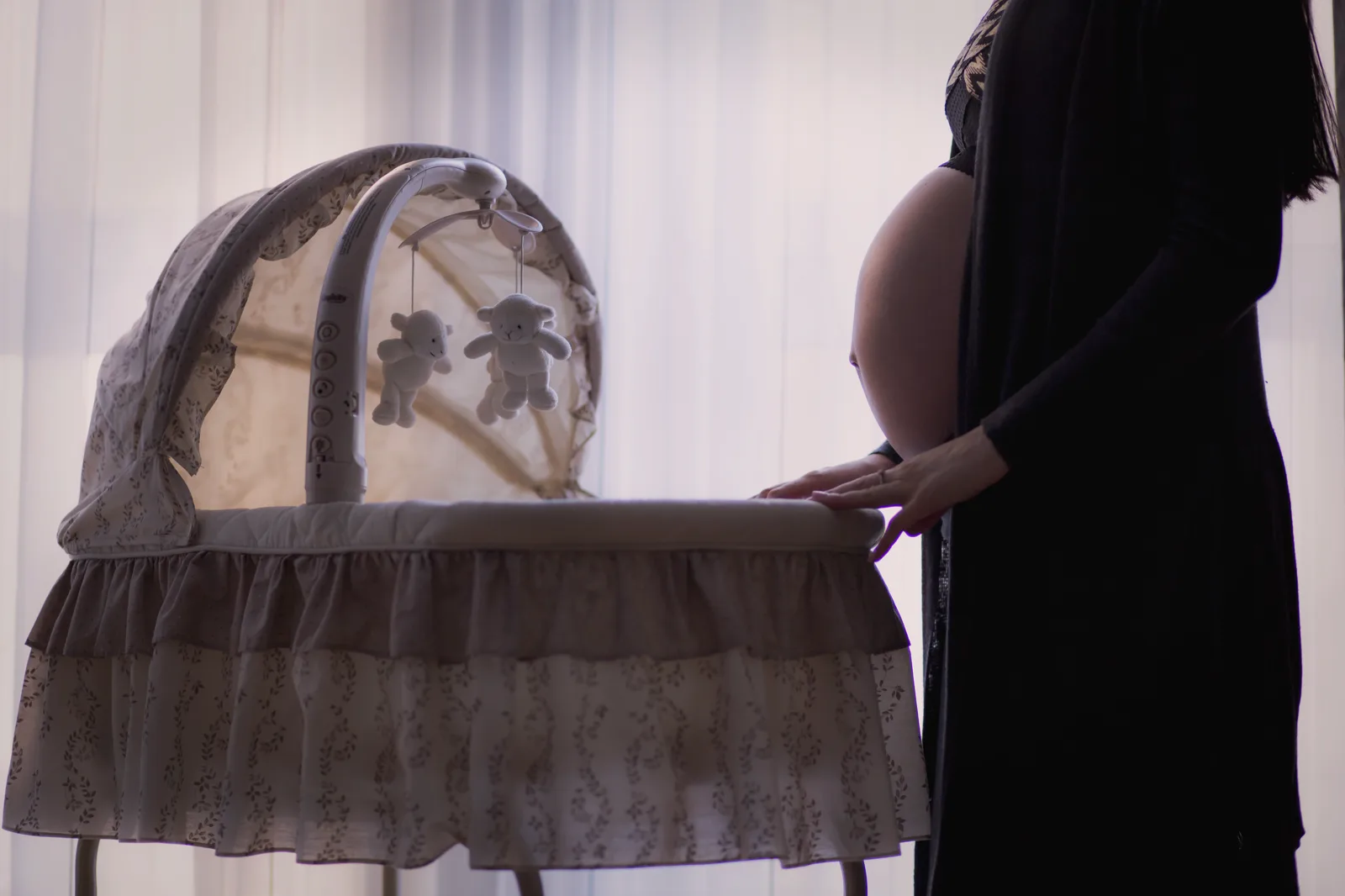 A pregnant woman in a long dark cardigan, seen from the shoulders down, rests her hands on the edge of a bassinet with a mobile of toy bears, in front of a bright curtained window