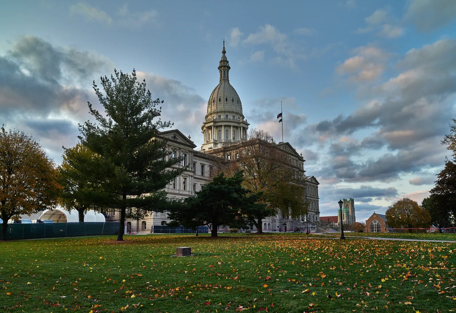 A white stone capitol building with a tall dome and two flags at half staff, seen across a wide lawn scattered with autumn leaves, with evergreen and leafy trees in front under a cloudy evening sky