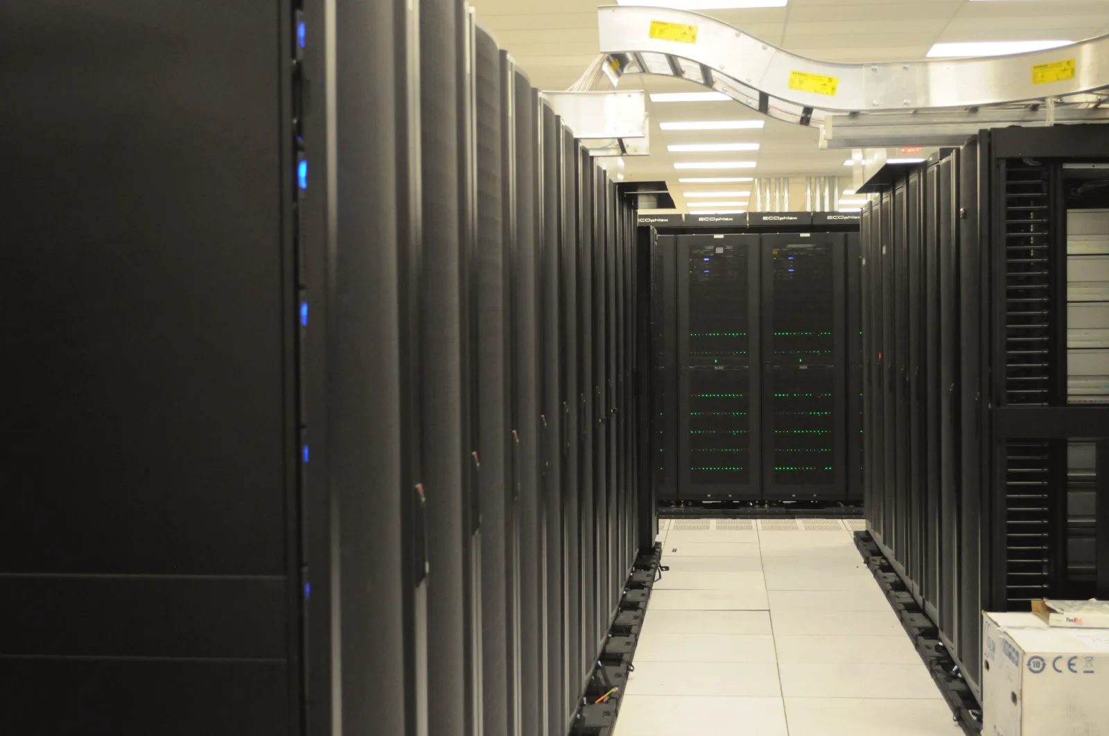 Two long rows of black server cabinets with small blue and green lights line an aisle with a white tiled floor, under a metal cable tray hanging from the ceiling