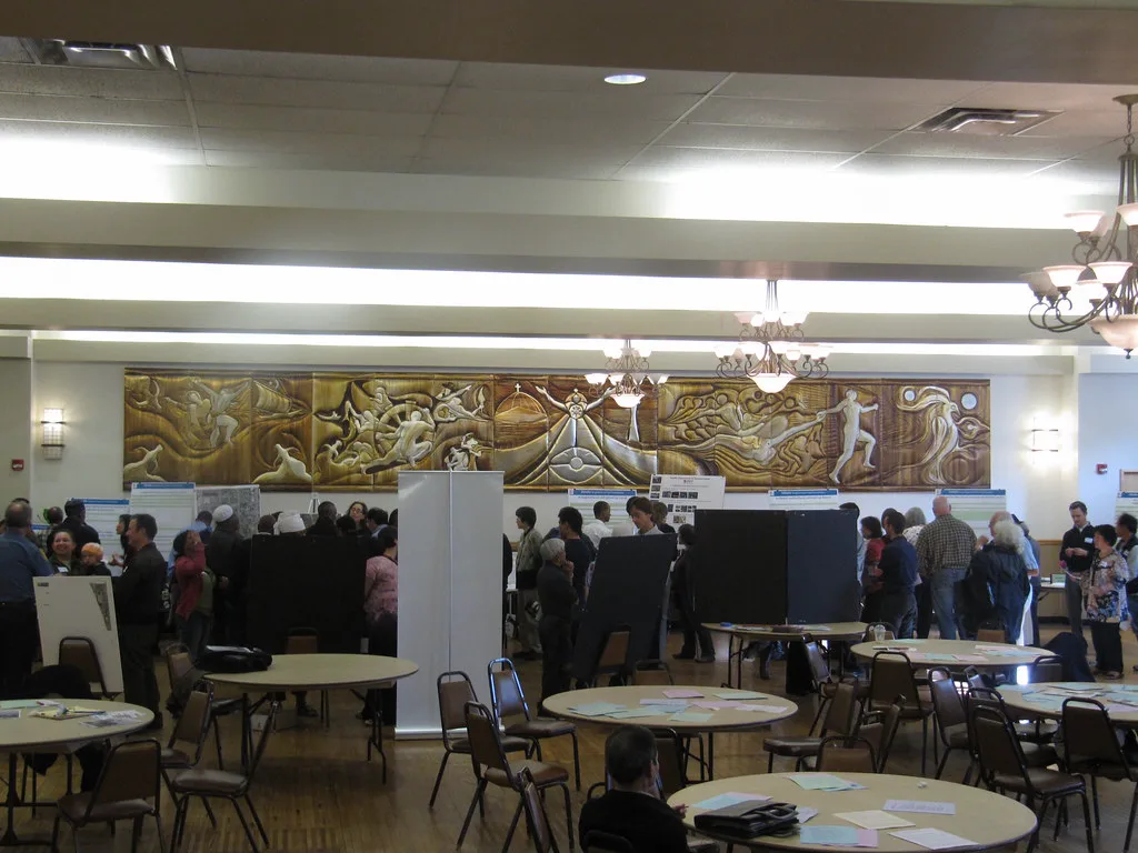 A large hall with a carved golden relief along the back wall, round tables with papers and folding chairs in front, and dozens of people standing among display boards in the distance