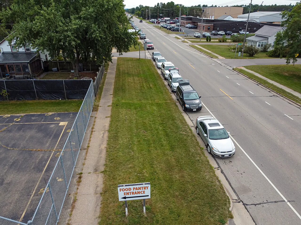 Seen from above, a line of cars waits along a street curb beside a grass strip with a sign reading Food Pantry Entrance