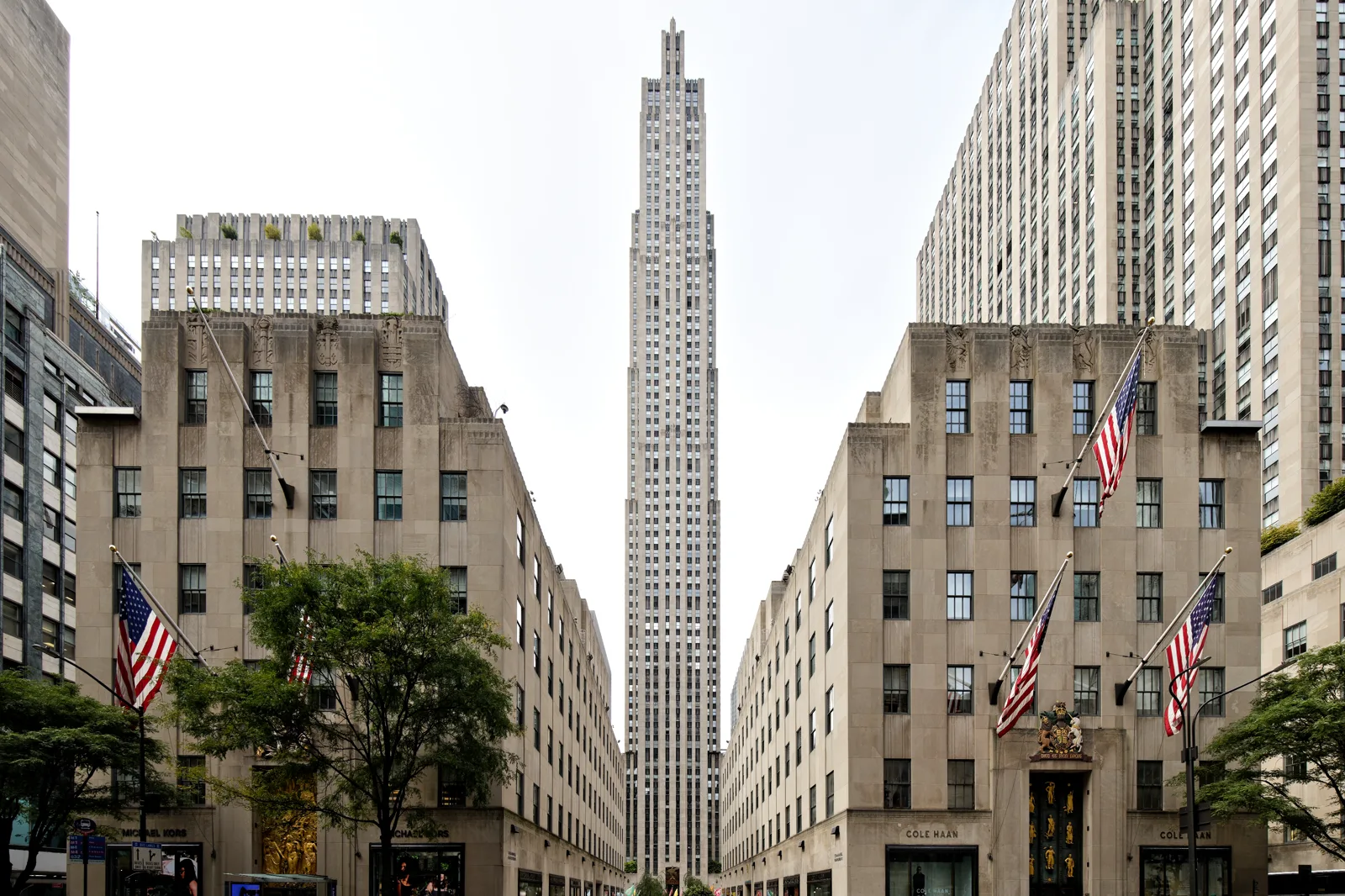 A slender limestone skyscraper rises at the end of a promenade between two low stone buildings flying American flags, with taller towers on either side under a pale sky