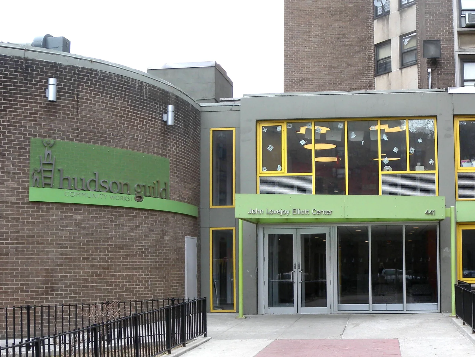 The entrance of a brick and gray community center building with yellow window frames, a green Hudson Guild sign on a curved brick wall and a green canopy reading John Lovejoy Elliott Center over glass doors